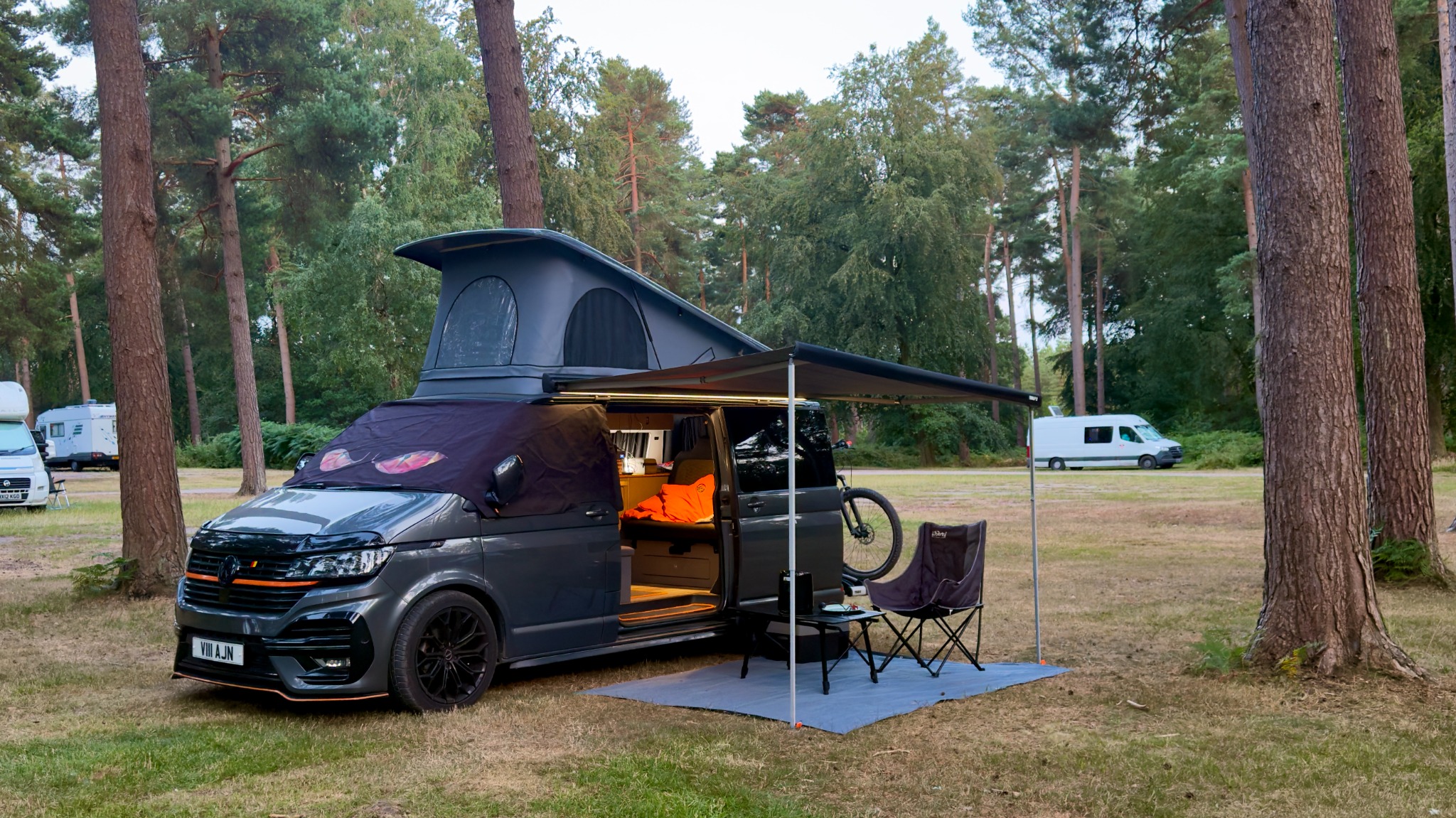 Vanilla set up among tall pine trees with the pop-top raised, side awning open and Torque carried at the rear